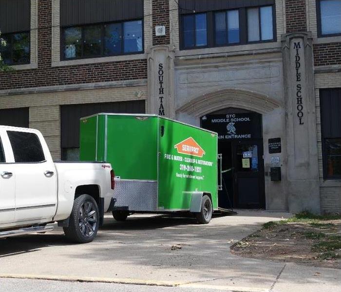 White pickup truck with green trailer in the back is in front of the entrance of a school.